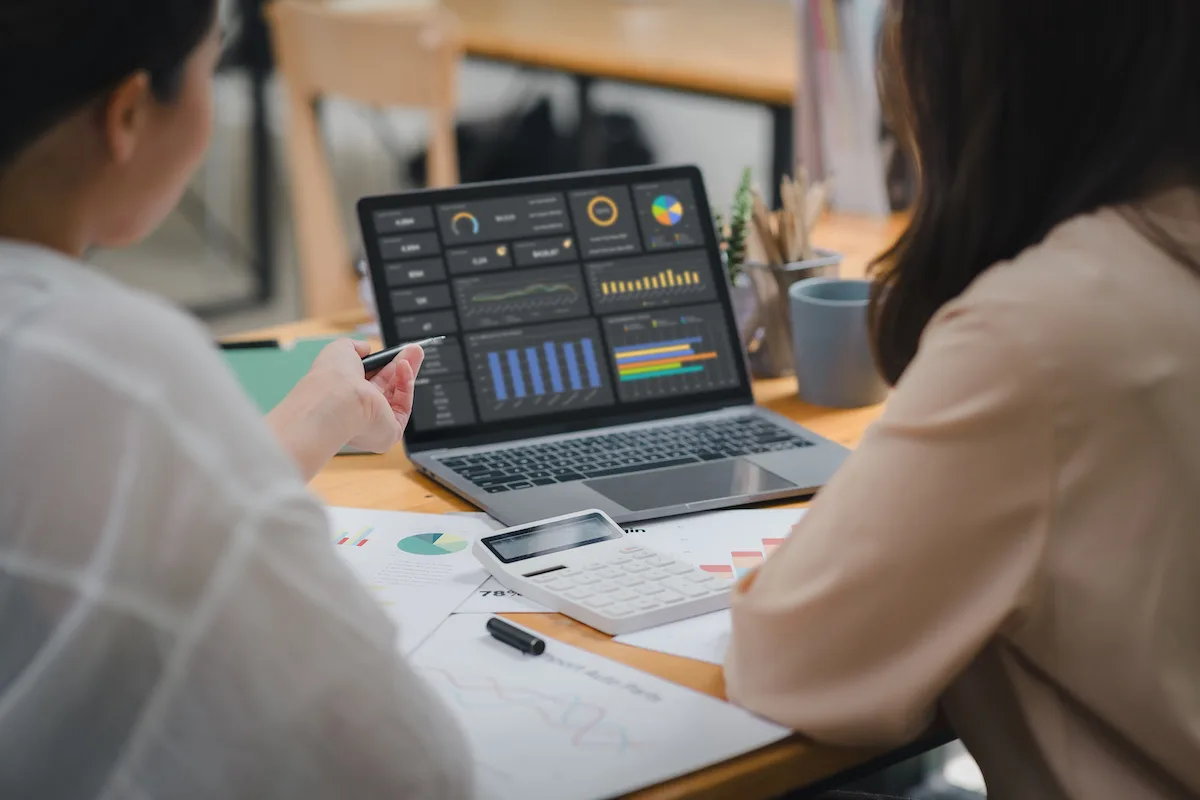 Two people sitting at a desk, pointing at a laptop screen displaying multiple analytics dashboards and charts.